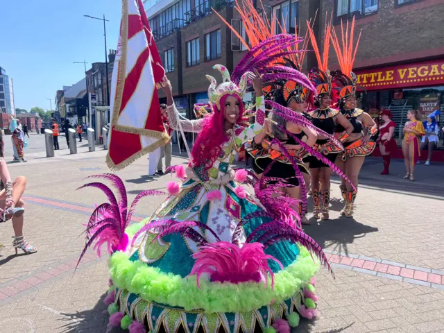 Amber Ogunsanya-William stands in her elaborate bright pink and green hooped costume, while bearing a flag.