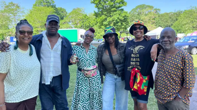 Six people with their arms around each other stand in a line and pose for a photo. They are smiling. There are trees, cars and stalls are behind them.