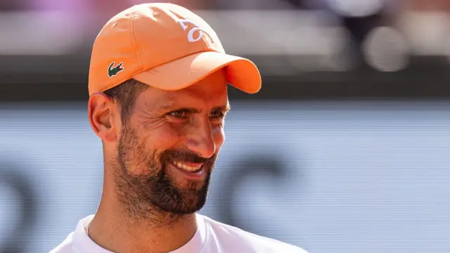 Head and shoulders of a smiling Novak Djokovic, wearing a white t-shirt, orange cap and dark beard