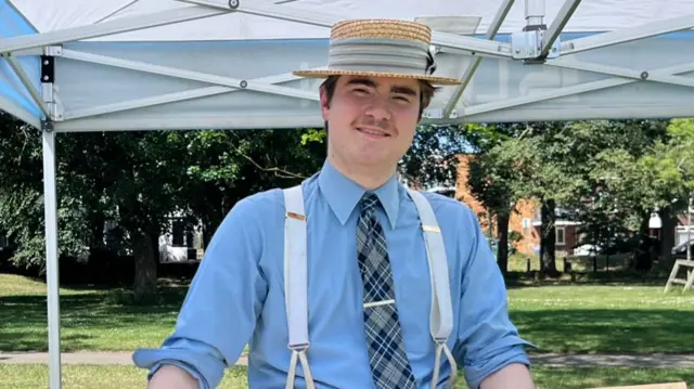 James Anderson has dark hair and is wearing a straw hat with a band of material around it. He is also wearing a blue shirt, a tartan tie and braces.