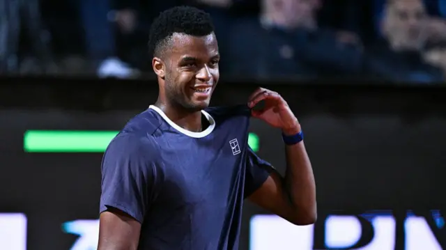Mpetshi Perricard, wearing a dark blue t-shirt, smiles and tugs at the left shoulder of his t-shirt with his left hand