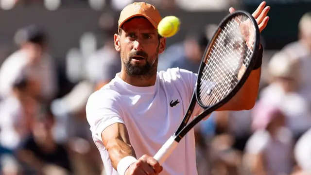 Novak Djokovic, wearing a white t-shirt and orange hat, plays a one-handed backhand with a black racquet in his right hand and the ball at eye level