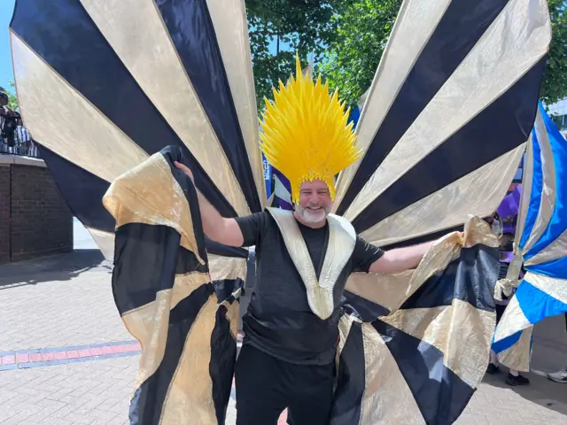 Andy Collins is holding on to huge black-and-gold striped wings and is wearing a pointy yellow headpiece.