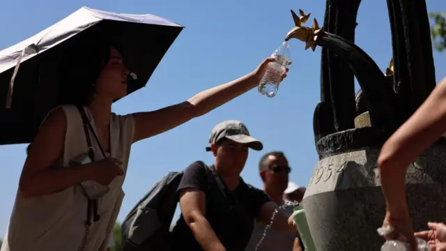 A woman fills a water bottle from a public tap while holding an umbrella to shield herself from the sun.