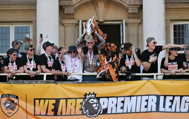 The players on the balcony cheer as a man wearing sunglasses with a medal around his neck lifts the trophy.