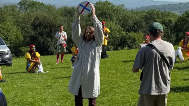 A man is holding up a cheese while dressed in a white robe. He is standing on grass and hills can be seen in the background.
