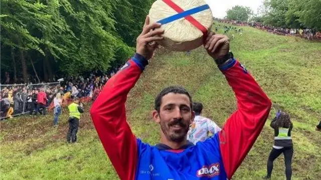 A man with a moustache is lifting a large wheel of Double Gloucester cheese. Behind him is a very steep hill.