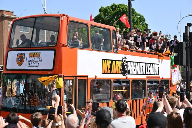 A close up of the players on the top of the open bus, some raise their drinks to cheer, others stand and laugh as the bus proceeds.