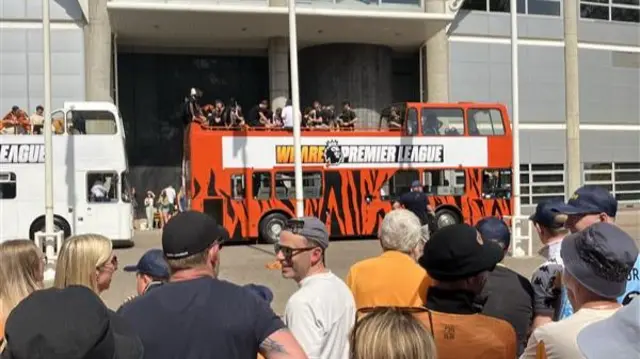 A small crowd watches a tiger-striped bus outside a football stadium.