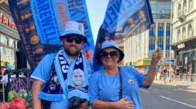 Manchester City fans Josh and Carla in the club kit waving flags ahead of the open top bus parade in Manchester