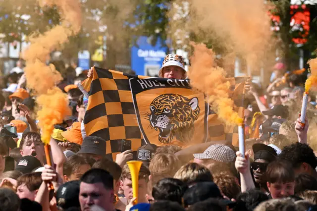 A young man sits on the shoulders of another and holds a Hull City flag up. orange flares and vuvuzelas can be seen in the crowd.