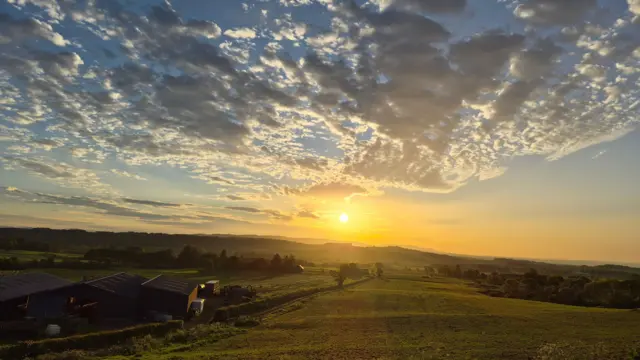 A sun rise across a farm with rolling hills far off. There's a barn in the bottom left