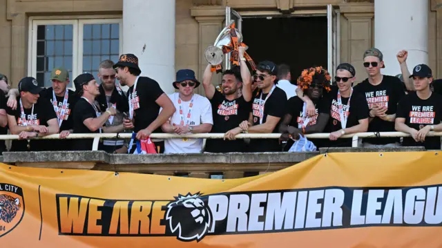 The players stand on a balcony and cheer, with one holding the Championship trophy.