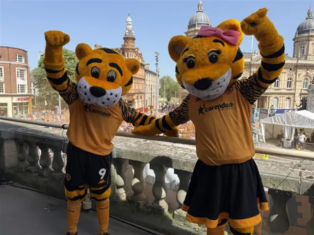 The Hull City Mascots, Roary and Amber, with furry tiger heads, cheer with their arms up on the City Hall balcony with the crowds in the background.