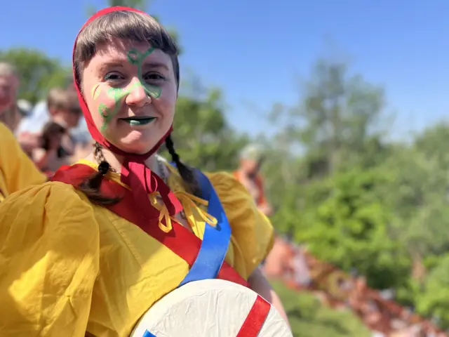 A woman is wearing face paint holding a wheel of cheese.