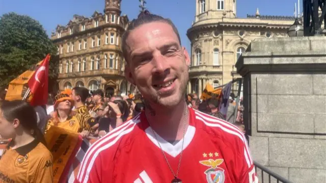 A man wearing a red football shirt smiles at the camera with crowds of Hull City fans behind him.