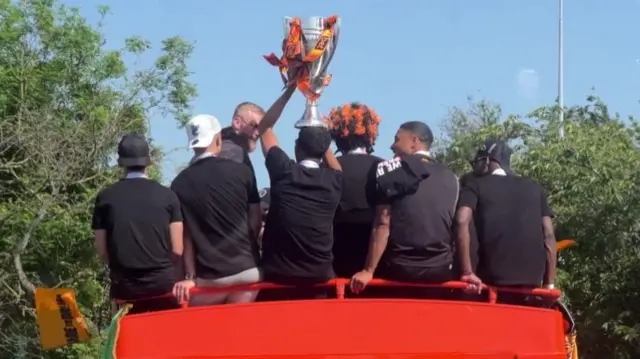 The players perch on the back of the open top bus, with the trophy balanced on the top of one of the player's heads. One of the other players wears a black and amber wig.