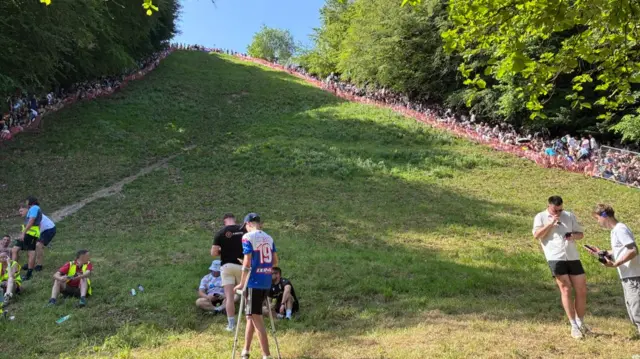 A few people are standing at the bottom of a very steep hill, which is covered largely by the shade.