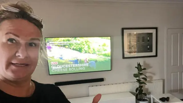 A woman standing in front of a TV with a BBC stream of the Gloucestershire cheese rolling. She has her thumbs up and is smiling at the camera.