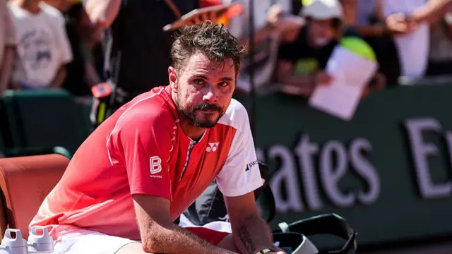 Stan Wawrinka, wearing an orange t-shirt and white shorts, sits in his chair at the side of the court looking emotional