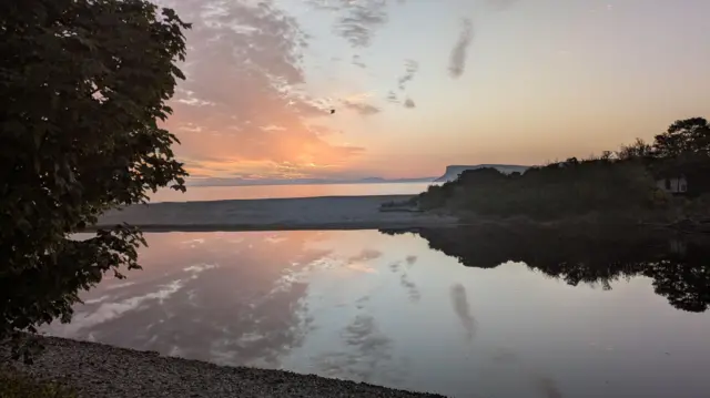 A sun rise above water, separated by a beach in between. A bird flies over head