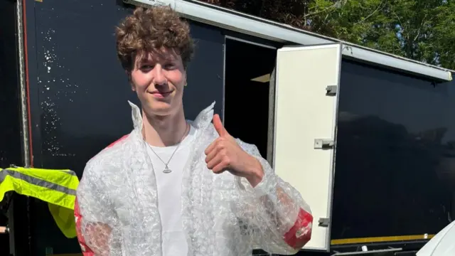 A man with a bubble wrap jacket gives a thumbs up. He has curly brown hair.