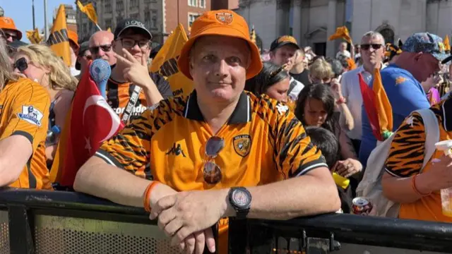 A man wearing a Hull City black and amber kit and hat stands with his hands over the barrier with crowds behind him.