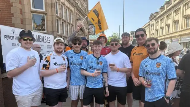 Ten men wearing Hull City shirts hold drinks and wave flags as they stand in the shade waiting for the parade to start.