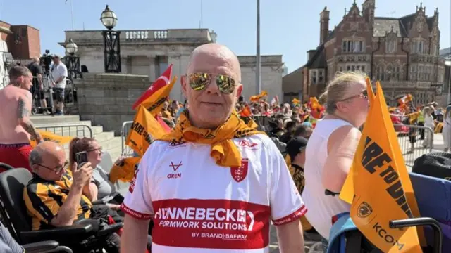 A man wearing a Hull KR rugby top and Hull City neckerchief and mirrored sunglasses stands in the crowd and smiles.