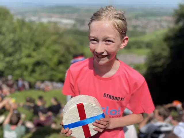 Dotty is standing on top of the hill in a pink T-Shirt holding up her cheese.