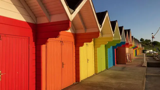 A row of bright coloured beach huts, red, orange, yellow, green and blue, going down a walkway far into the distance