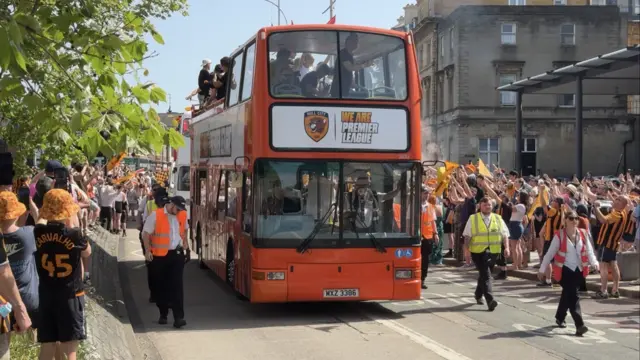 A red open-top bus travels down a road surrounded by football fans who are dressed in amber and black and celebrating. A sign on the bus reads: "We are Premier League".