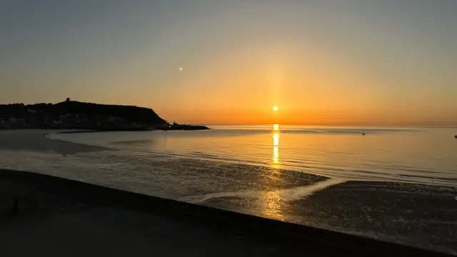 Sun rises over a calm sea. There's an empty beach and a hill in the distance