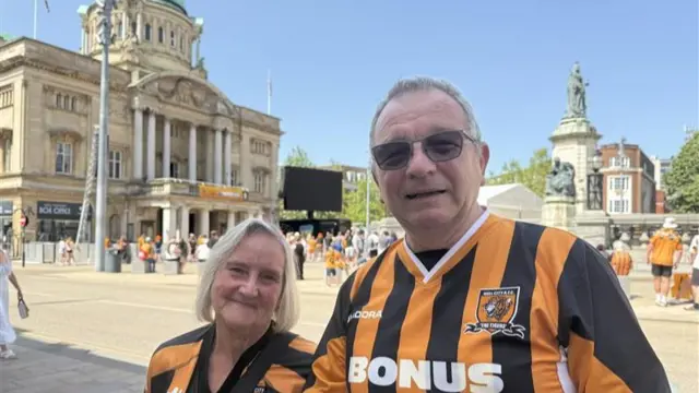 A man and a woman wearing black and amber Hull City shirts stand in the town centre in the sunshine. The man wears sunglasses.