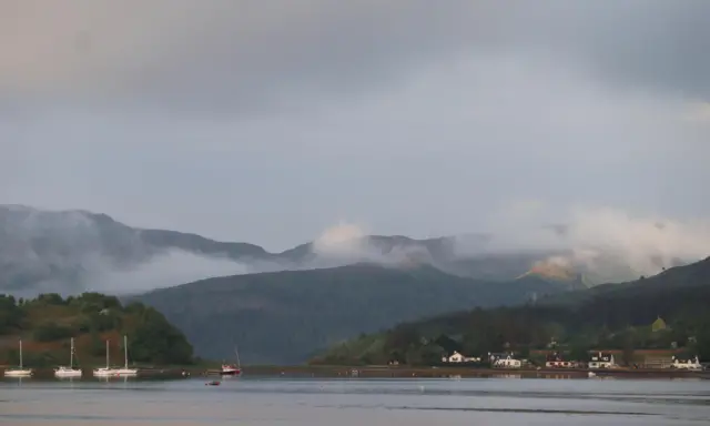 A sun rise over misty mountains that have clouds draped over them. There is the sea in the foreground, with a small village on its shores and some sail boats anchored. it's very still