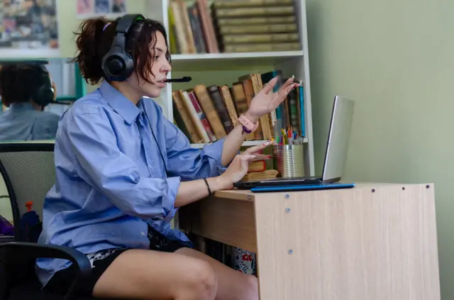 A woman on a video call wearing a blue shirt, black shorts and a headset