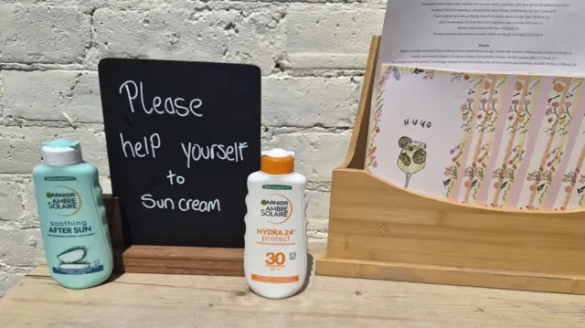 Two bottles of suncream on a table with a chalk sign saying: "Please help yourself to suncream."