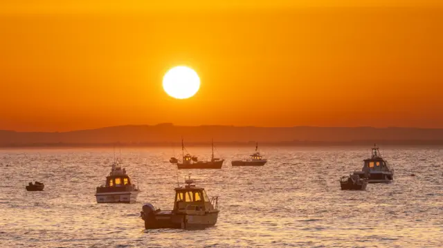 Boats moored in the sea which is slightly choppy as the sun rises, glowing the entire sky orange and reflecting the sea