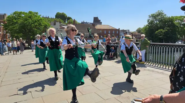 A troupe of eight clog dancers on a paved river bridge. They are wearing calf-length green skirts, black waistcoats and white, short-sleeved collared shirts. They are jigging and holding tassels. People are watching.