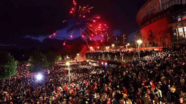 A huge crowd outside the stadium, filling up every bit of space. Fireworks go off, casting a red glow