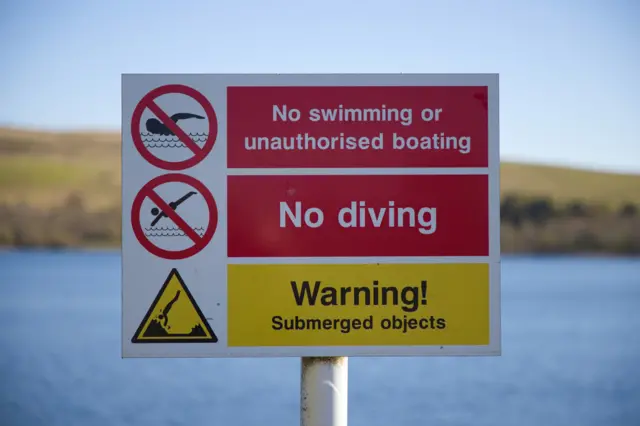 A red and yellow safety placed in front of a UK reservoir warning against swimming and diving due to hidden submerged objects