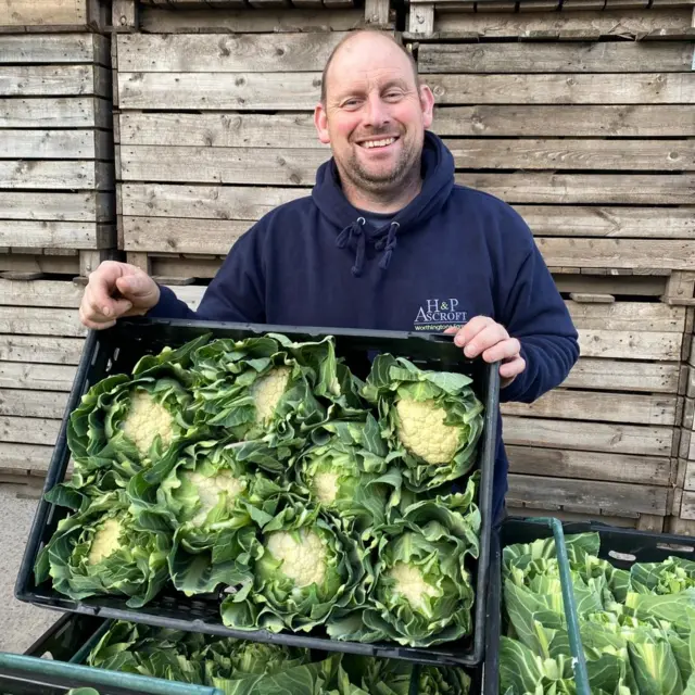 Jon Forshaw holding crate of freshly harvested cauliflower heads in front of stacked wooden crates outdoors.