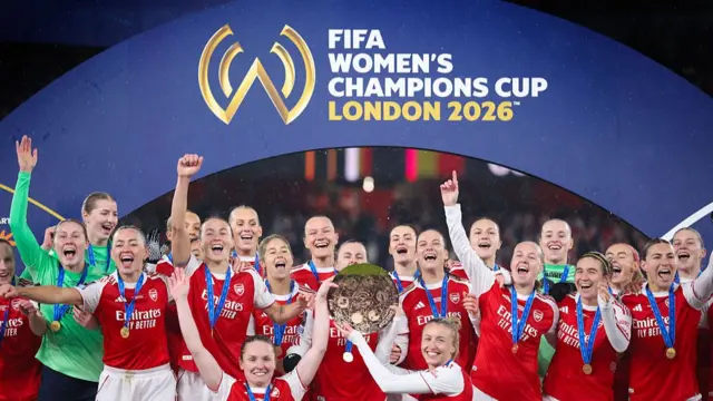 The Arsenal women's team standing cheering holding a trophy in the middle under a banner calling them champions