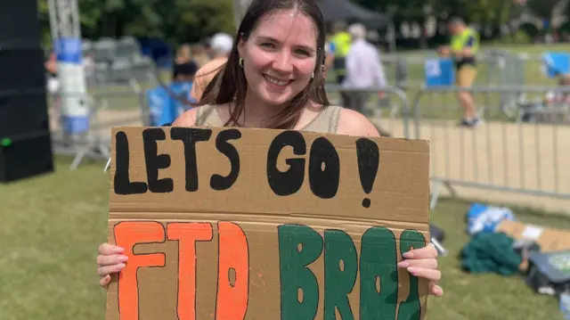 Brown haired woman holding up cardboard sign. The sign says Lets Go FTD Bros!