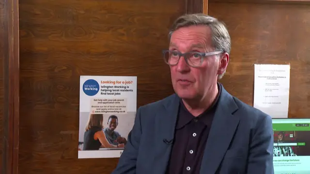 Alan Milburn sits down during a broadcast interview. He is wearing a dark blue suit with a darker shirt underneath