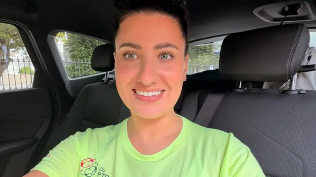 A woman looks into the camera with a broad smile. She is sitting in the back seat of a car, and the seats and grey upholstery are visible. She is wearing a light-green running top. She has black curly hair tied up.