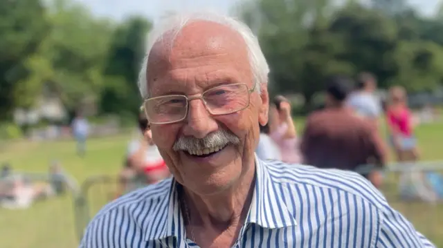 John is smiling at the camera. He is wearing glasses and has a thick moustache. He has white hair and is wearing a navy and white striped shirt.