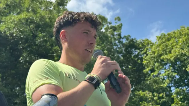 a shot from below of a man speaking into a microphone. The sky behind him is blue with hardly any clouds