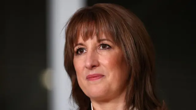 Rachel Reeves looks on, with a black and blurred white background behind her.