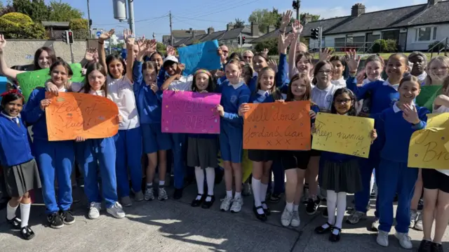 Around 30 pupils wave toi the camera with multiple different signs with messages of support. They are dressed in Blue and White uniform, smiling and waving at the camera.